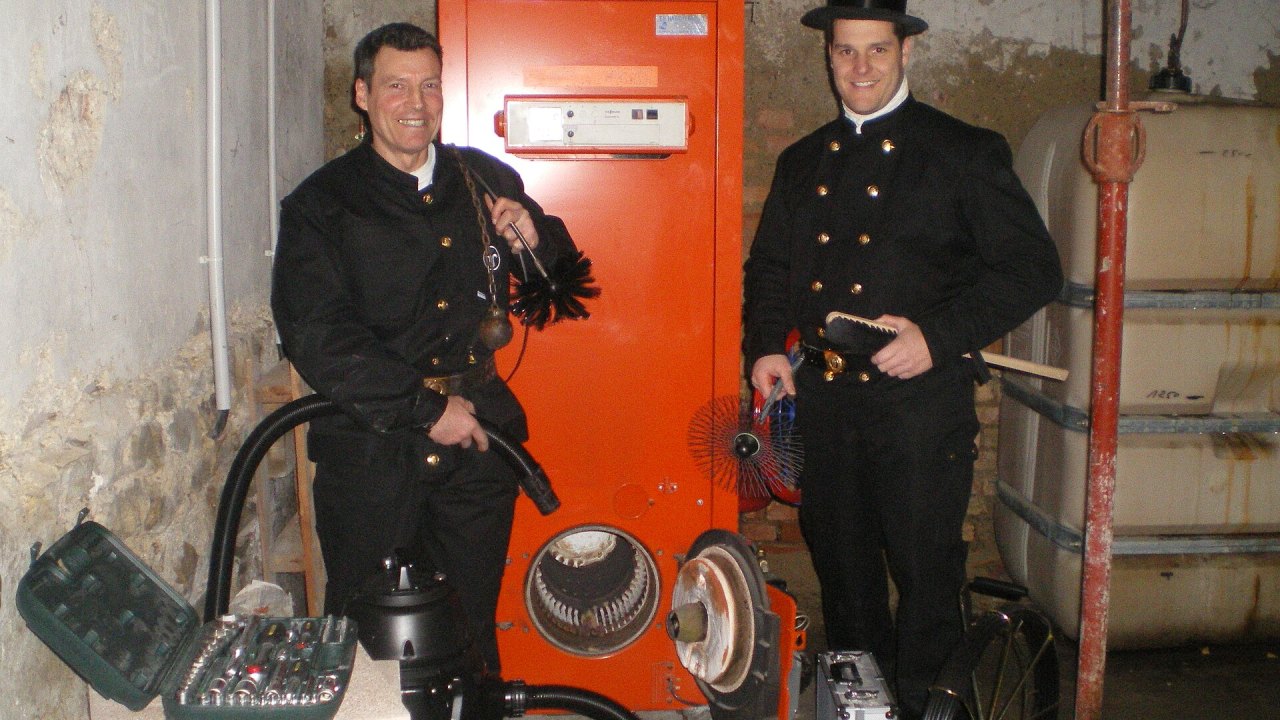 Due spazzacamini con scovoli, aste, aspiratore e attrezzi accanto a una caldaia in un locale tecnico
