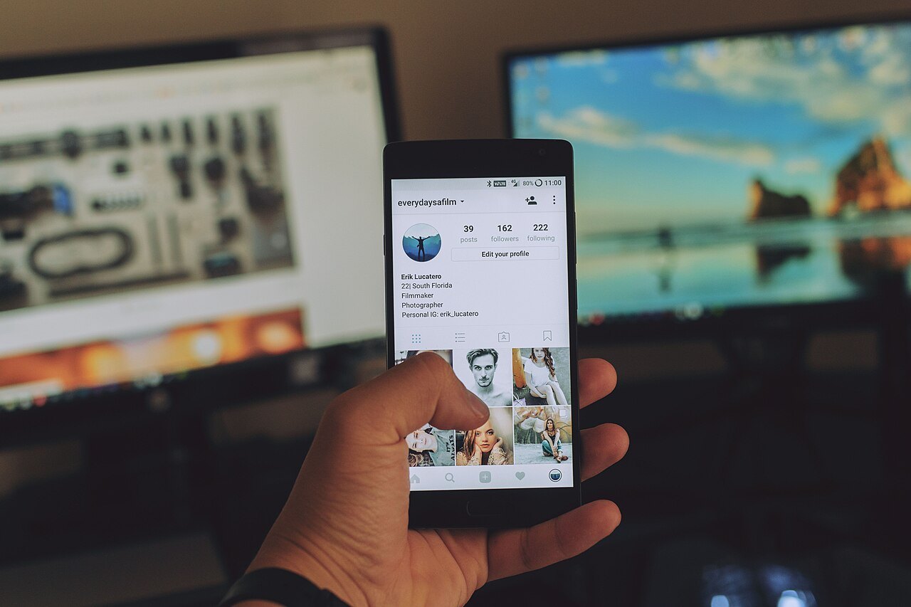 Mano che tiene un telefono con un profilo Instagram aperto davanti a due monitor