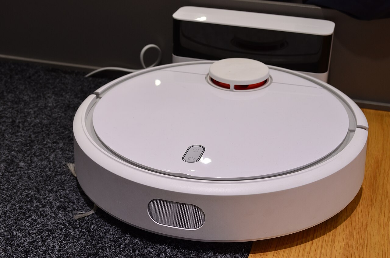 Robot aspirapolvere con torretta LiDAR davanti alla sua base di ricarica, fra tappeto e parquet
